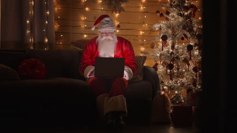 Gesamtplan-Vorderansicht-Ein-Echter-Weihnachtsmann-Arbeitet-Nachts-Mit-Einem-Laptop-Mit-Brille-Im-Licht-Der-Weihnachtsbeleuchtung-Auf-Dem-Hintergrund-Eines-Weihnachtsbaums.-Heimarbeit